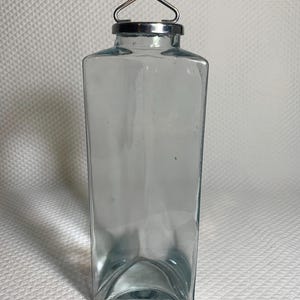 May include: A clear, rectangular glass bottle with a silver metal rim and a triangular handle. The bottle has a slightly curved base and a narrow, elongated shape. The glass has a slight blue tint.
