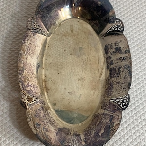 May include: An oval silver-toned metal tray featuring a scalloped edge with intricate cut-out detailing. The surface displays significant dark tarnish and patina throughout, with a smooth, recessed center and decorative embossed floral motifs.