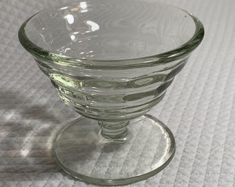 May include: A clear glass dessert dish with a round bowl and a stem. The bowl has a series of horizontal ridges. The dish sits on a circular base. The glass is transparent.