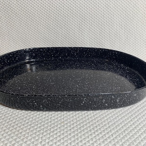 May include: An oval-shaped black enamel roasting pan featuring a speckled white pattern throughout the surface. The pan includes two curved metal handles attached to each side, providing a functional design for lifting and carrying the vessel.