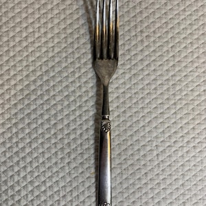 May include: A silver-toned metal dinner fork featuring four slender tines and a straight handle. The handle is decorated with a small, embossed shell-like motif at both the top and the bottom, providing a subtle textured detail to the flat surface.