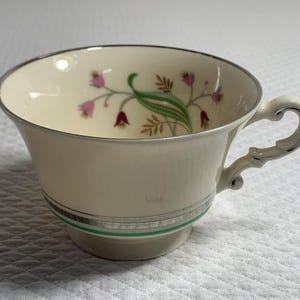 May include: Cream-colored teacup with a silver rim and handle. The cup features a floral design with pink and green accents. A silver band and a green stripe encircle the base of the cup. The cup is on a white surface.
