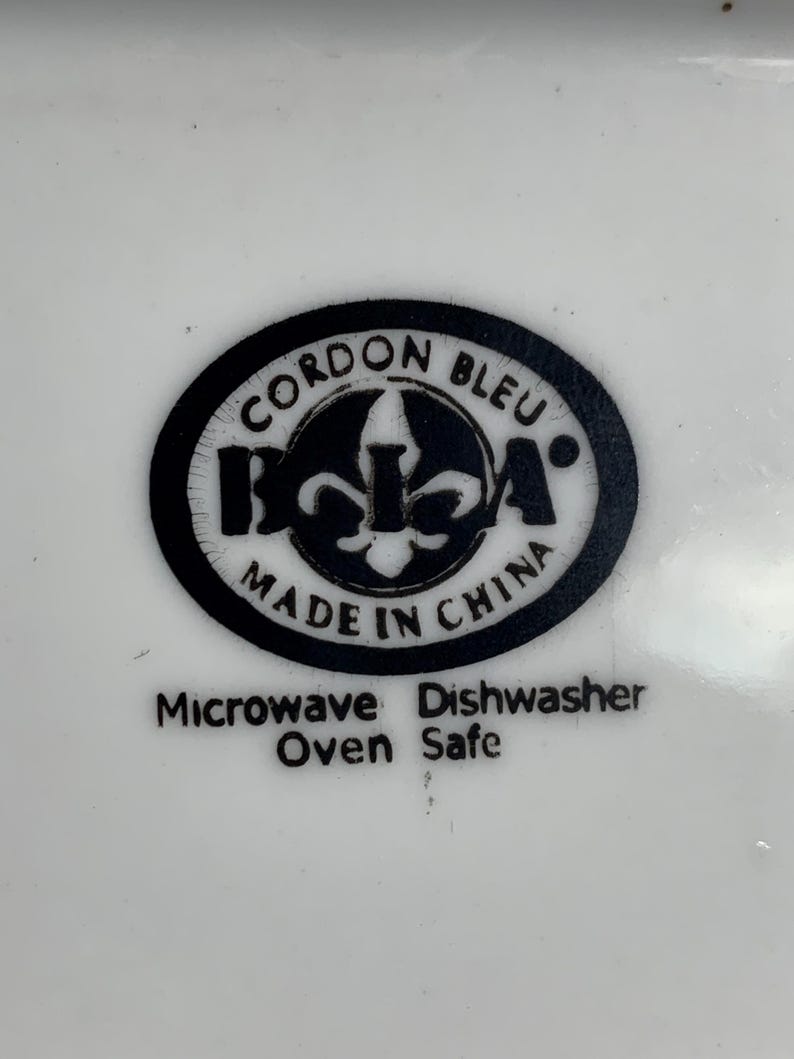 May include: A white surface with a black circular logo reading "CORDON BLEU" and "BCA" with a fleur-de-lis symbol. Below the logo, it says "MADE IN CHINA". The text "Microwave Oven Safe" and "Dishwasher Safe" is also visible.