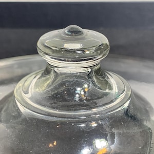 May include: Close-up of a clear glass lid with a rounded knob. The lid has a slightly domed shape and sits atop a clear glass dish. The glass has a few imperfections, adding to its vintage charm. The background is a dark gray.