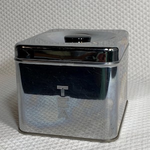 May include: A square, chrome-finished metal tea canister with a reflective surface. The word TEA is embossed vertically on the front side in a stylized font. The canister features a removable lid with a small, round, black knob on top.