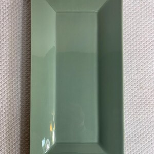 May include: A rectangular, sage green ceramic serving platter. The platter has a slightly raised, angled rim and a glossy finish. The shape is ideal for serving appetizers or small dishes.