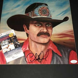 May include: A signed album cover featuring a portrait of a man wearing a brown cowboy hat with colorful embellishments. The man has a mustache and is wearing a red shirt. The background is a sunset landscape.