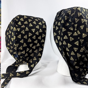 May include: Black scrub hat with a gold and black pattern of the Deathly Hallows symbol. The hat has a tie that can be tied at the back of the head.
