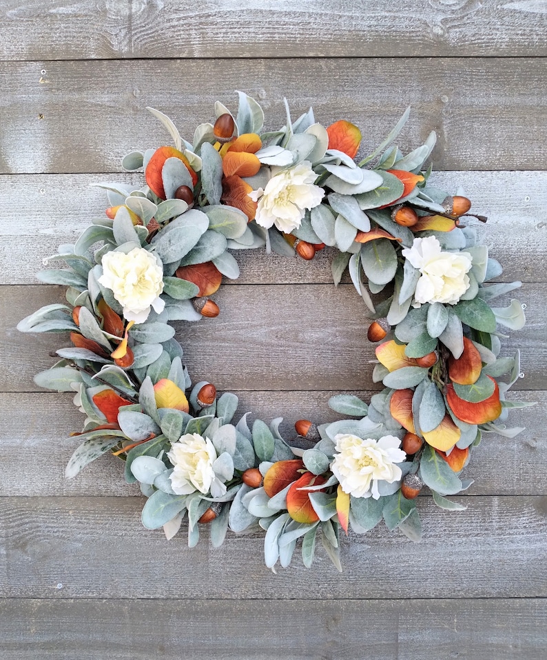 May include: A fall-themed wreath with white flowers, orange and yellow leaves, and brown acorns. The wreath is made of artificial greenery and is displayed against a gray wooden background.