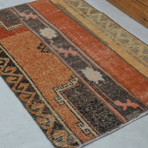 May include: A rectangular rug with a geometric pattern in shades of orange, brown, and beige. The rug has a fringed edge and is made of a woven material.