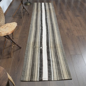 Pode incluir: Um tapete comprido e estreito com listras e uma faixa branca no centro. O tapete é feito de tecido tecido e tem um aspecto desgastado. O tapete está sobre um piso de madeira.