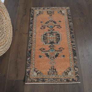 May include: A rectangular, orange-toned rug with a central geometric design in black, teal, and beige. The rug has a patterned border and is placed on a dark wood floor. A woven basket is visible in the upper left corner.