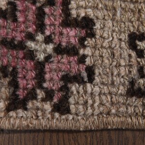 Op de afbeelding: Close-up van een deel van een geweven tapijt met een bloemenpatroon. Het tapijt heeft een beige achtergrond met donkerbruine en roze bloemmotieven. De randen hebben franjes.