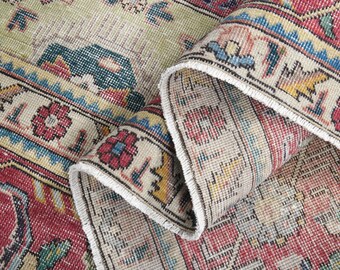 May include: Close-up of a section of a vintage rug with a folded edge. The rug features a complex pattern with red, cream, blue, and yellow hues. The design includes floral and geometric elements, showcasing a distressed, antique aesthetic.