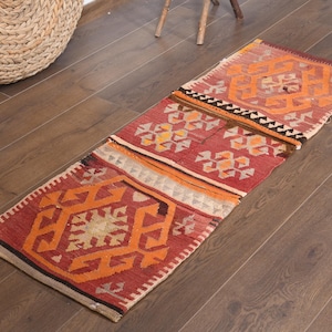 Puede incluir: Una alfombra rectangular con un patrón rojo y naranja. La alfombra presenta diseños geométricos y está colocada sobre un suelo de madera. Una cesta tejida es visible en el fondo.