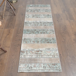 May include: Rectangular rug with a distressed, vintage appearance. The rug features alternating bands of beige and faded teal with floral and geometric patterns. The rug is placed on a wooden floor, showcasing its design and texture.