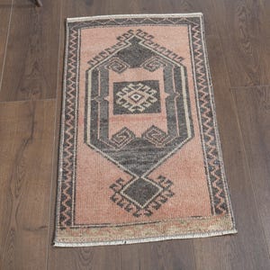 May include: A small, rectangular rug with a faded pink field and a central geometric design in grey and black. The rug features a patterned border and is placed on a wooden floor.