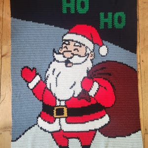 May include: A rectangular crochet blanket featuring a graphic of Santa Claus wearing a red suit with a white trim, a black belt with a gold buckle, and black boots. Santa carries a large maroon sack. The text HO HO HO is in green.