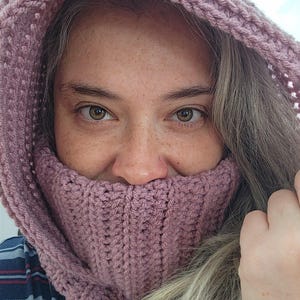 Op de afbeelding: Een stofroze gehaakte capuchon en col. De capuchon is omhoog getrokken en bedekt het hoofd en de nek, waarbij het colgedeelte het onderste deel van het gezicht bedekt. De haaksteek creëert een geribbelde textuur. De persoon draagt een blauw en grijs geruit overhemd.