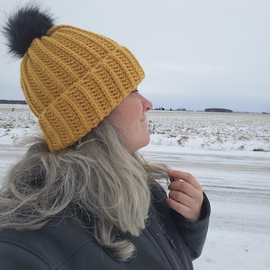 May include: Mustard yellow knitted beanie hat with a black faux fur pom-pom. The hat features a ribbed design. A warm accessory for cold weather, the beanie is shown in a winter setting.