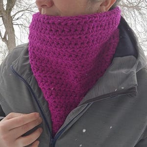 May include: A magenta crochet cowl, worn around the neck. The cowl is made of thick yarn, creating a textured appearance. The person is wearing a grey jacket with a zip. Snowflakes are visible in the background.