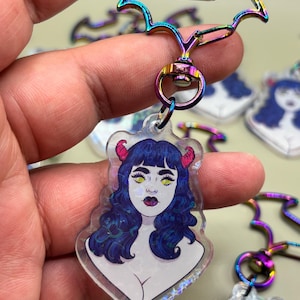 May include: A keychain featuring a devil-themed illustration. The charm has a clear, iridescent background with a cartoon image of a woman with blue hair, red horns, and yellow eyes. The keychain hardware is a rainbow-colored bat shape.