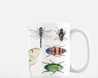 Puede incluir: Taza de cerámica blanca con una ilustración colorida de varios insectos, incluyendo una libélula, una abeja, una polilla y escarabajos. Los insectos se representan en un estilo realista con detalles intrincados.