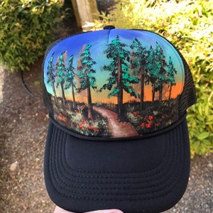 May include: Black trucker hat with a scenic landscape design. The hat features a black brim and mesh back. The front panel displays a painted scene of a path through a forest with trees and a colorful sunset.