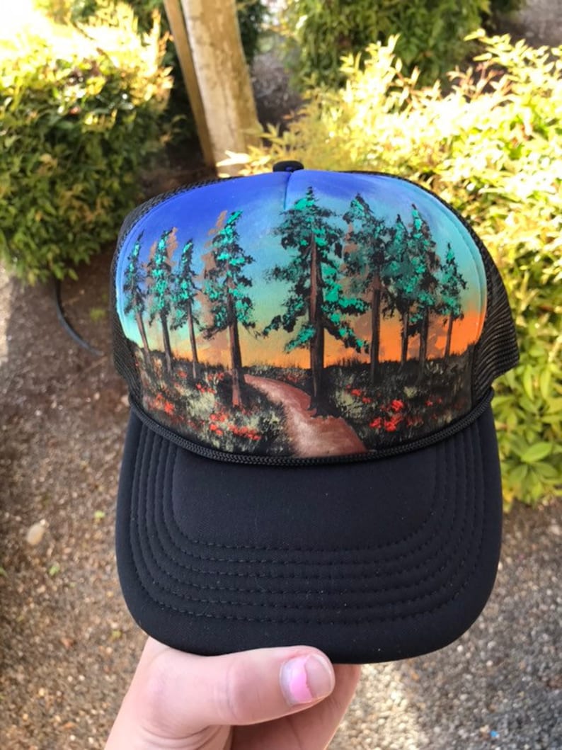 May include: Black trucker hat with a scenic landscape design. The hat features a black brim and mesh back. The front panel displays a painted scene of a path through a forest with trees and a colorful sunset.