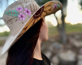 May include: A beige cowboy hat with a painted sunflower and pink flowers. The hat is decorated with green leaves and has the text "Bittersweet Canvas" on the brim.