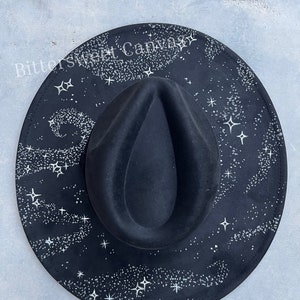 May include: Black fedora hat with a celestial design. The hat features a black crown and brim, with a white starry night sky pattern printed on the brim. The text "Bittersweet Canvas" is printed on the brim.