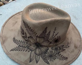 May include: A tan fedora hat with a wide brim, featuring a detailed floral design in dark brown ink. The hat has the text "Bittersweet Canvas" on the crown. The hat is made of a soft, suede-like material.