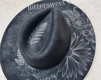 May include: A black felt fedora hat with a white floral design painted on the brim. The design features leaves, ferns, and flowers.