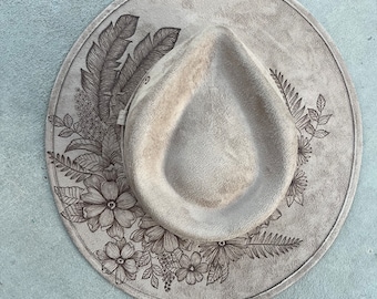May include: A beige fedora-style hat with a wide brim. The brim is decorated with a floral and leaf design. The hat has a teardrop-shaped crown and a band around the base of the crown.