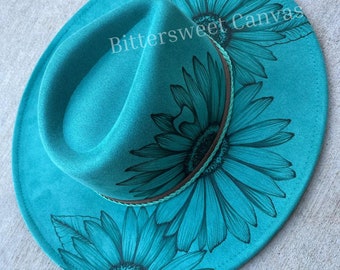 May include: Teal felt fedora hat with a brown band and two hand-painted sunflowers in a dark teal color. The hat has the text "Bittersweet Canvas" written on it.