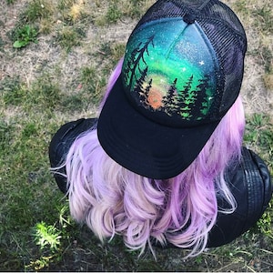 May include: A black mesh trucker hat with a hand-painted design of a forest scene with a green and blue aurora borealis in the background.