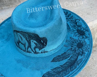May include: A teal blue fedora hat with a black buffalo silhouette, feathers, and floral designs. The hat is decorated with the text "Bittersweet Canvas".