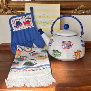 May include: A white enamel teapot with a blue handle and lid, decorated with colorful teacup designs. A blue oven mitt and a small tea towel with teapot patterns are also present. A cookbook is in the background.