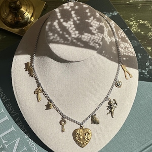 Op de afbeelding: Een zilveren ketting met goudkleurige bedels, waaronder een hartvormige hanger, een sleutel, een kat en een engel. De ketting wordt getoond op een neutraal gekleurde buste, met een boek op de achtergrond.
