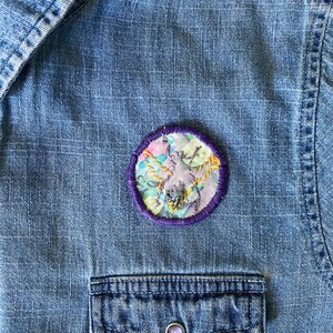 K&ouml;nnte beinhalten: Nahaufnahme eines Jeanshemdes mit einem runden, gestickten Aufn&auml;her. Der Aufn&auml;her zeigt ein florales Design mit lila, rosa und gelben Akzenten, umgeben von einem lila Rand. Das Hemd hat eine Brusttasche mit einem Knopf.