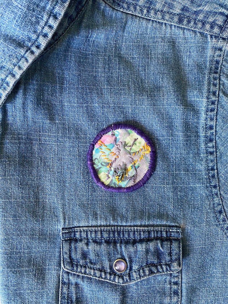 K&ouml;nnte beinhalten: Nahaufnahme eines Jeanshemdes mit einem runden, gestickten Aufn&auml;her. Der Aufn&auml;her zeigt ein florales Design mit lila, rosa und gelben Akzenten, umgeben von einem lila Rand. Das Hemd hat eine Brusttasche mit einem Knopf.