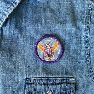 K&ouml;nnte beinhalten: Ein runder, gestickter Aufn&auml;her mit einem stilisierten Motten-Design in Lila, Orange und Wei&szlig; auf hellblauem Hintergrund. Der Aufn&auml;her hat einen lila gestickten Rand und ist auf einem blauen Jeanshemd befestigt.