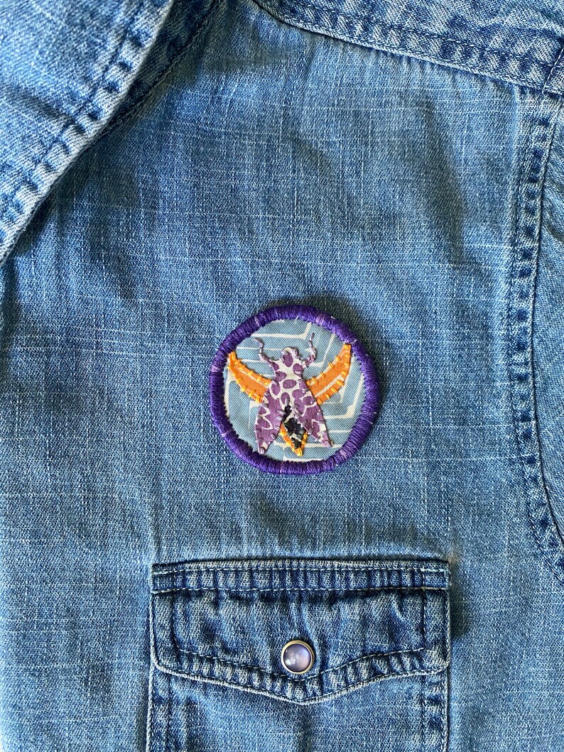 K&ouml;nnte beinhalten: Ein runder, gestickter Aufn&auml;her mit einem stilisierten Motten-Design in Lila, Orange und Wei&szlig; auf hellblauem Hintergrund. Der Aufn&auml;her hat einen lila gestickten Rand und ist auf einem blauen Jeanshemd befestigt.