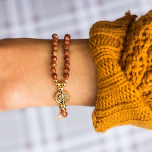May include: A gold and brown beaded bracelet with a gold pendant featuring a religious emblem. The bracelet is made of small, faceted, reddish-brown beads. The pendant is round with intricate detailing. The bracelet is worn on a wrist, with a mustard-colored sweater sleeve in the background.
