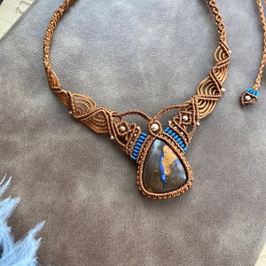 May include: A brown macrame necklace with a blue and brown teardrop-shaped opal pendant. The necklace is accented with small silver beads.