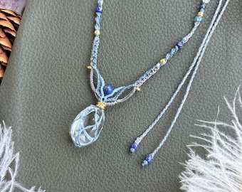 May include: A macrame necklace featuring a clear, teardrop-shaped crystal pendant encased in light blue cord. The necklace includes small lapis lazuli beads, clear crystal accents, and gold-toned metal spacers along the light blue cord.