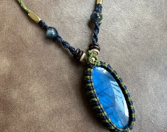 Puede incluir: Un collar artesanal con un gran colgante ovalado de piedra preciosa azul. El colgante está encerrado en un diseño de macramé con cordón verde y azul marino. El collar incluye detalles en latón y pequeñas cuentas.
