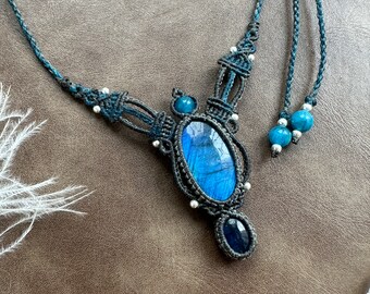 Puede incluir: Un collar de macramé azul oscuro y plateado con un gran colgante de labradorita ovalado y pequeños detalles de piedras azules.