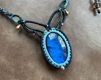 Puede incluir: Un collar con un colgante de labradorita azul, cordón de macramé negro y verde. El collar presenta cuentas doradas y un diseño anudado.