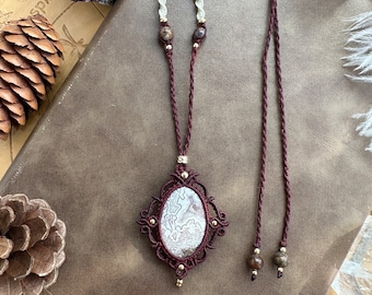 Könnte beinhalten: Eine braune Schnurkette mit einem weißen Achat-Anhänger in einer Makramee-Umrandung. Der Anhänger ist von goldenen Perlen umgeben. Die Kette liegt auf einer braunen Lederoberfläche.